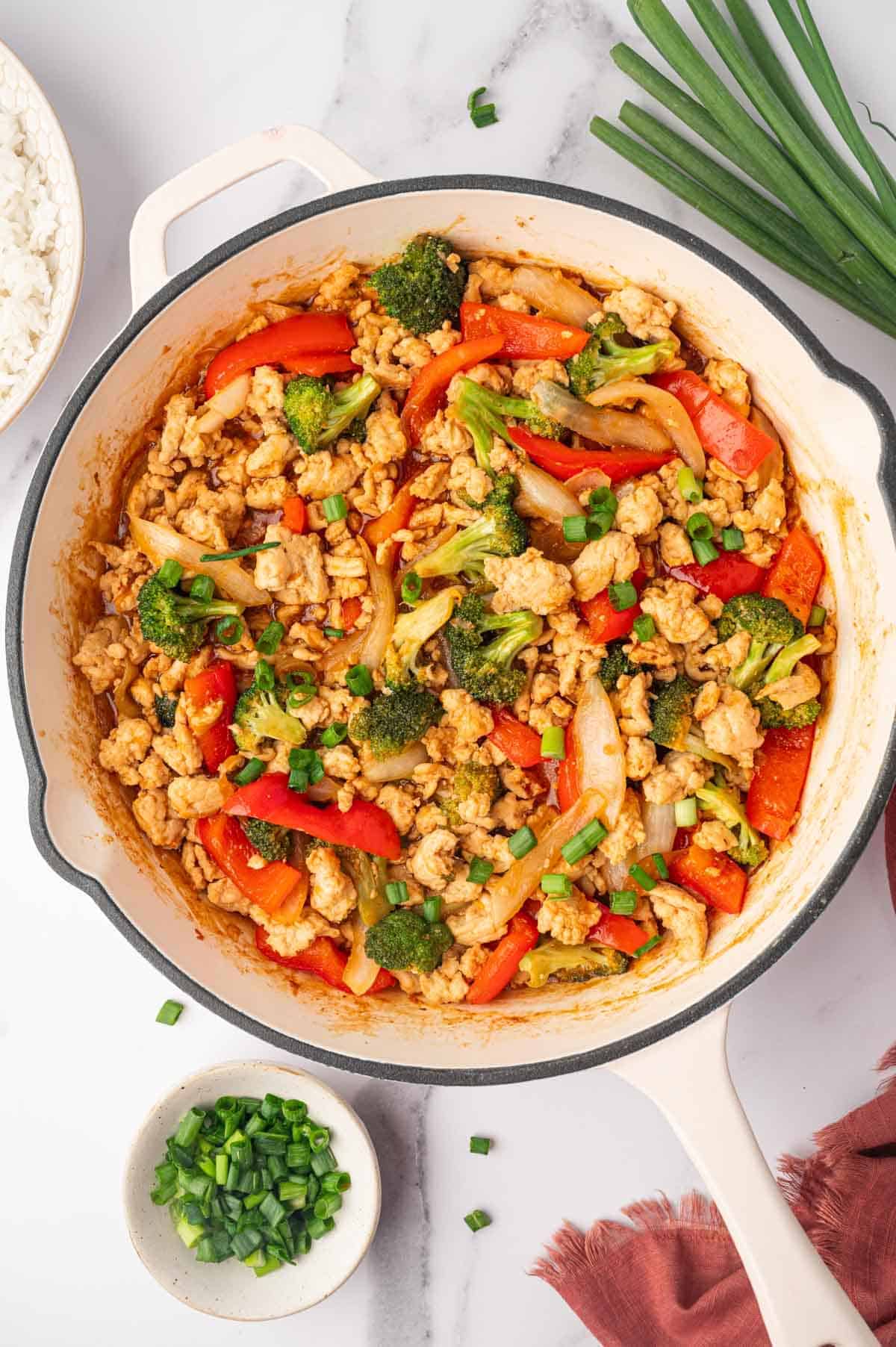 A skillet filled with stir-fried chicken, broccoli, red bell peppers, and onions in a savory sauce, garnished with chopped green onions. A bowl of rice and extra green onions are nearby on a white surface.