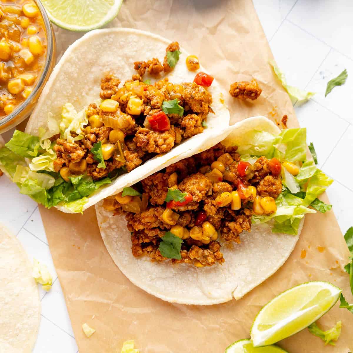 Two soft corn tortillas filled with seasoned ground meat, corn, red pepper, lettuce, and cilantro, served on parchment paper with lime wedges on the side.