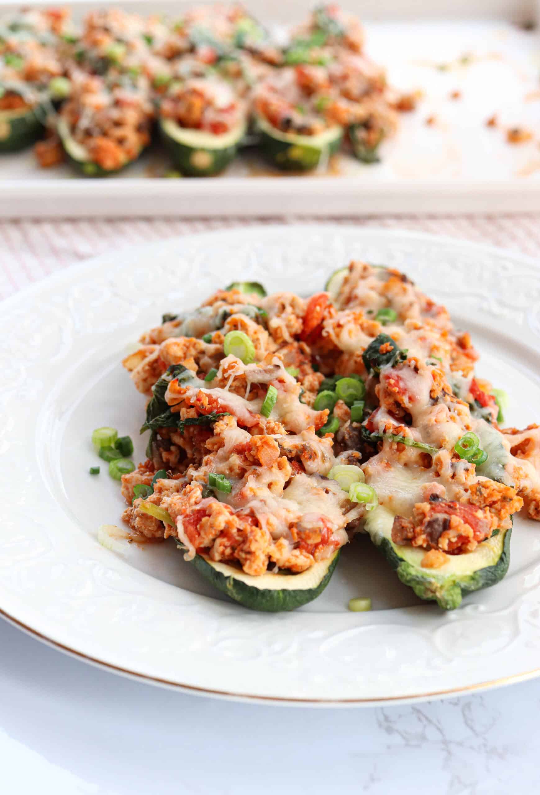 Two zucchini halves stuffed with a mixture of ground meat, vegetables, tomato sauce, and melted cheese, garnished with chopped green onions, served on a white plate. More stuffed zucchini are visible in the background.