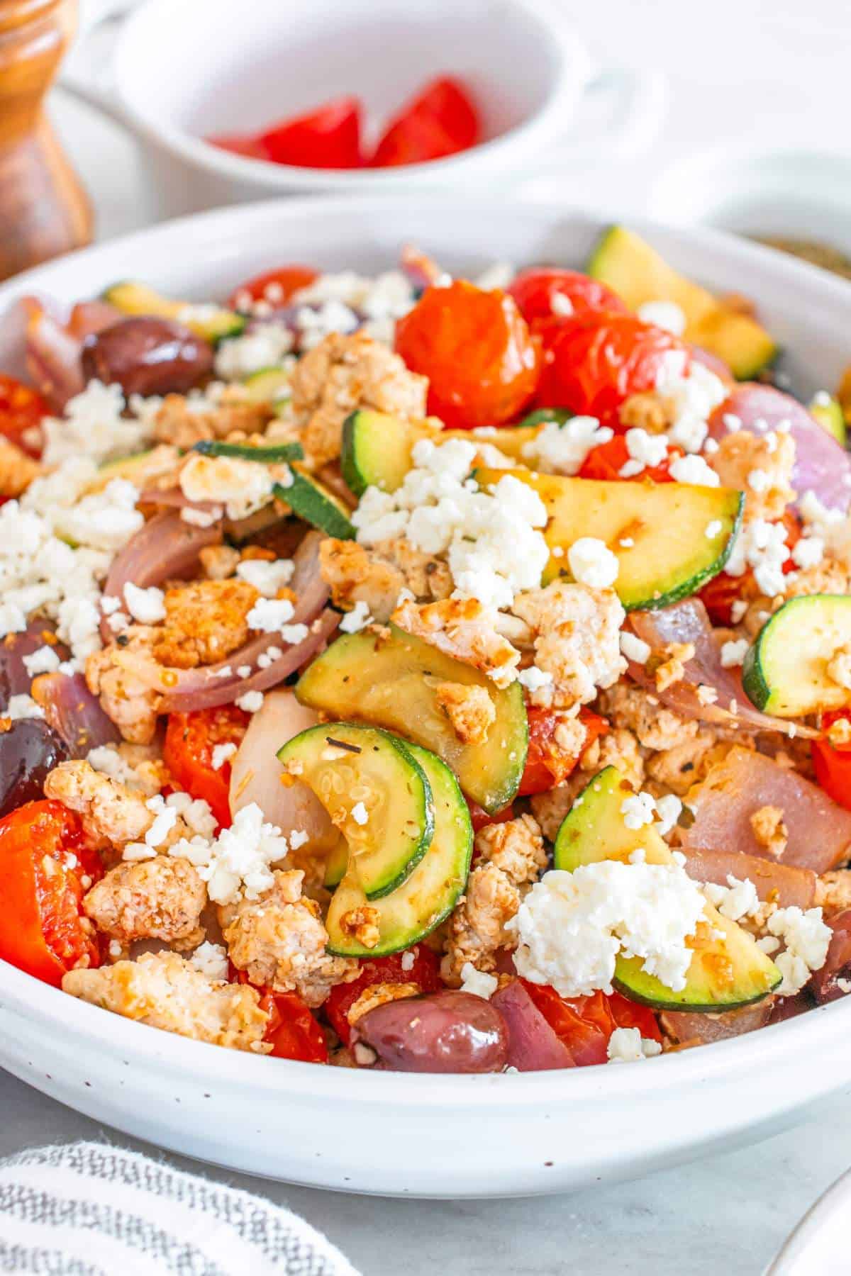 A close-up of a colorful Mediterranean salad with crumbled feta cheese, zucchini slices, cherry tomatoes, red onions, kalamata olives, and ground turkey in a white bowl.