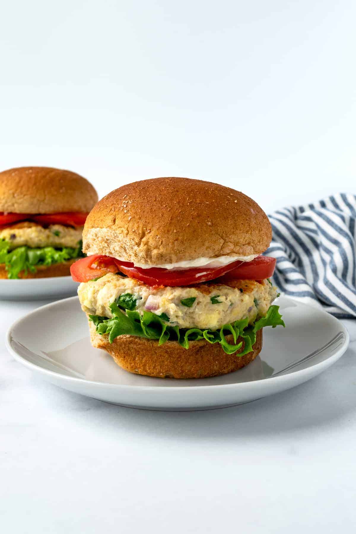 A chicken burger with lettuce, tomato slices, and mayonnaise on a whole wheat bun sits on a white plate, with another burger and a striped cloth in the background.