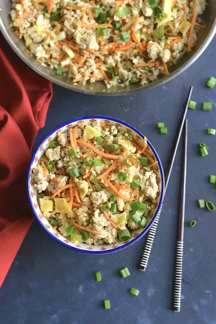 A bowl of fried rice with scrambled eggs, ground meat, sliced carrots, and chopped green onions sits next to a pair of chopsticks on a dark surface, with more fried rice and a red cloth in the background.