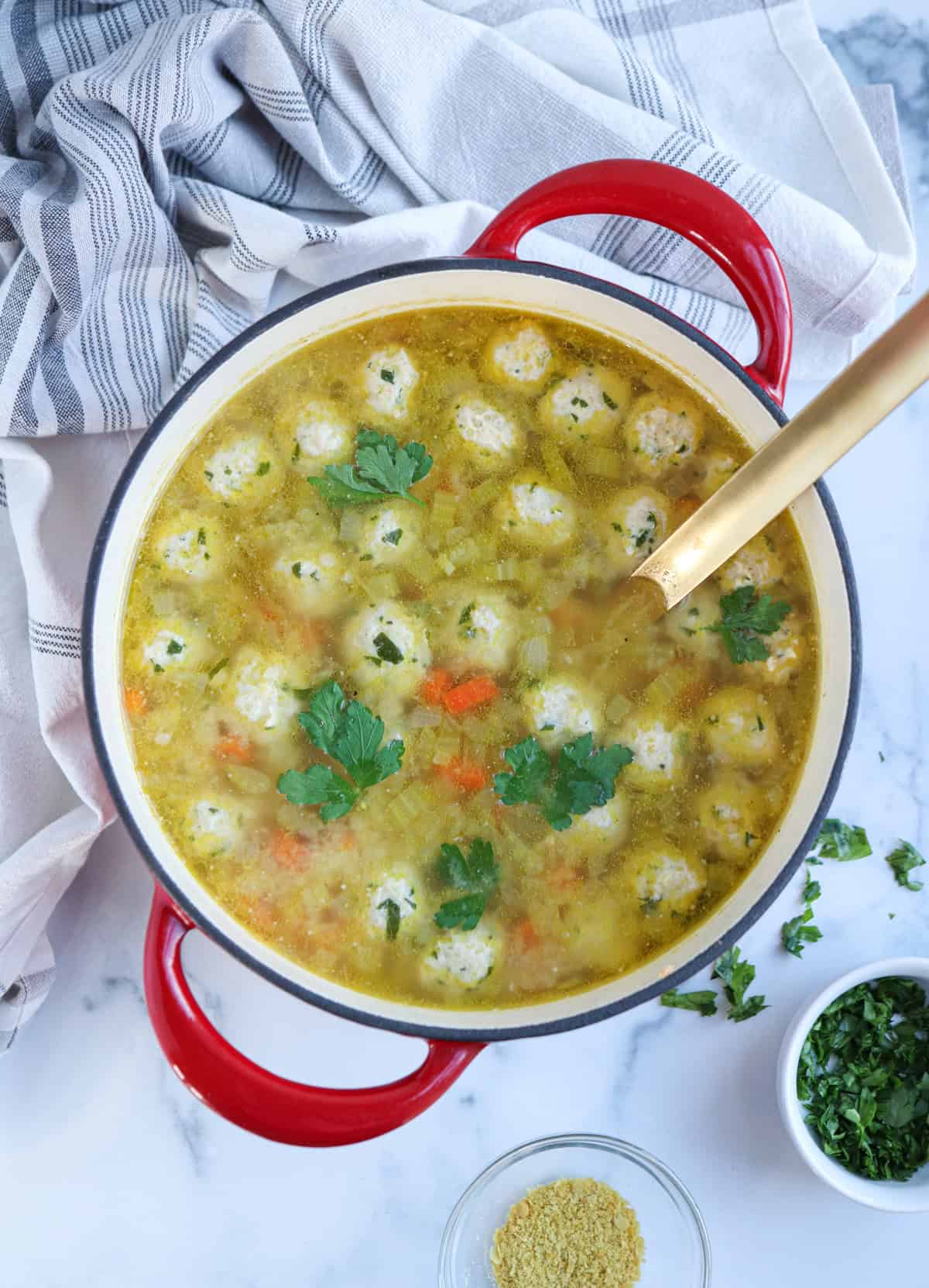 A red pot filled with soup containing small dumplings, chopped vegetables, and garnished with fresh parsley. A gold ladle rests inside the pot, with a striped towel and small bowl of greens nearby.
