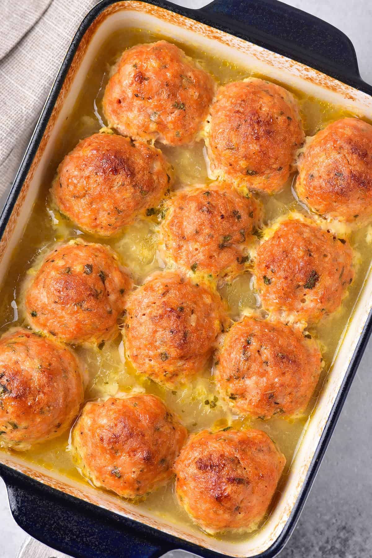 A baking dish filled with golden-brown meatballs in a light, herb-seasoned broth, freshly baked and arranged neatly in rows.