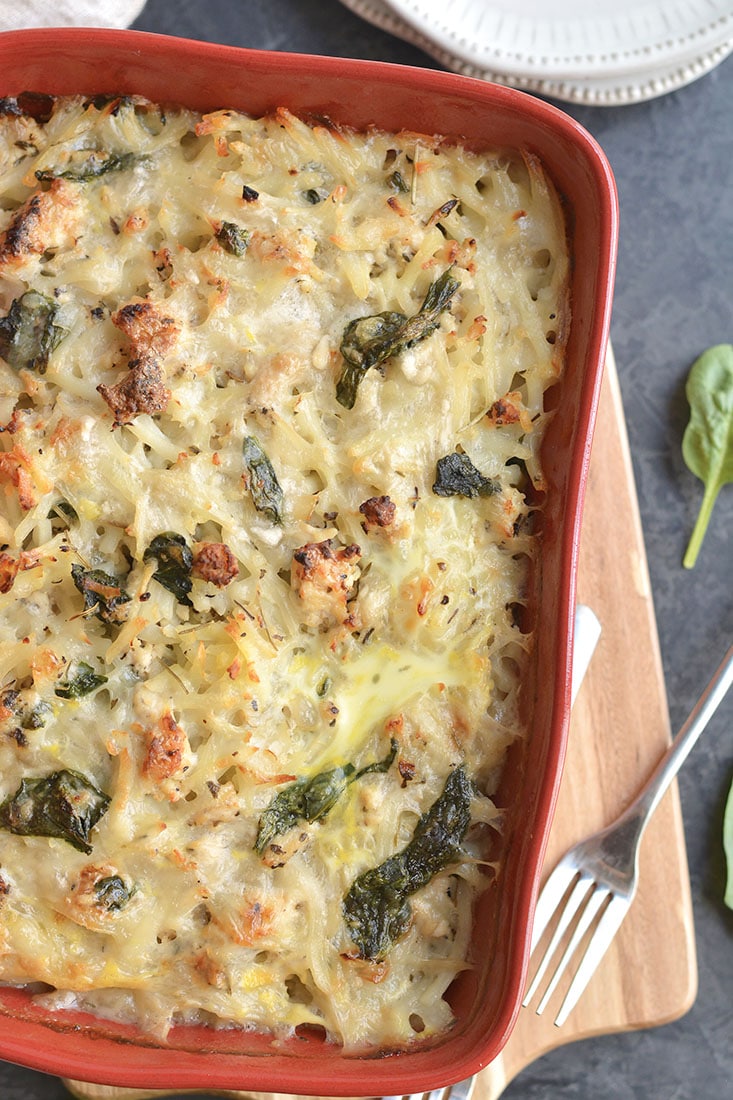 A baked pasta casserole with melted cheese, spinach, and golden brown edges in a red dish on a wooden board, next to a fork and white plate. Fresh spinach leaves are on the side.