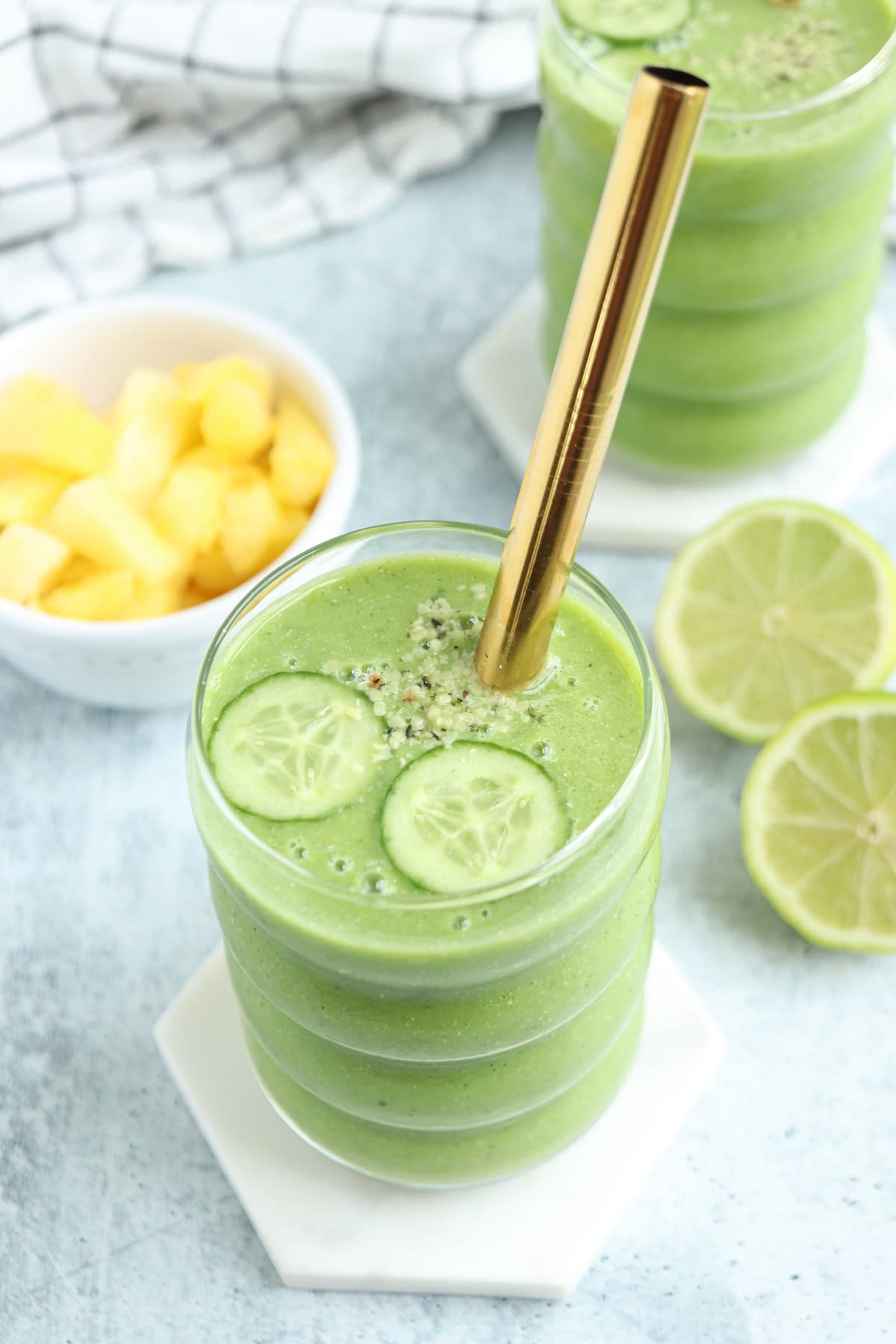 A pineapple cucumber smoothie in a glass with a gold straw, garnished with two cucumber slices and seeds. Fresh lime slices and a bowl of pineapple pieces sit nearby on a light blue surface.