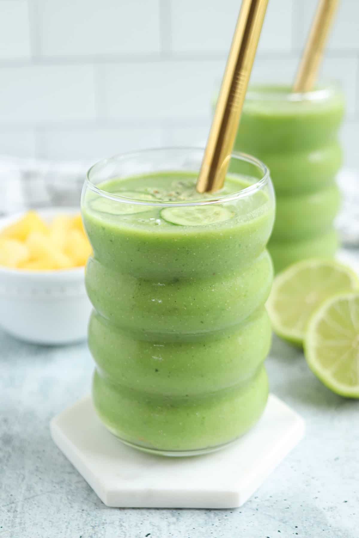 A glass of green smoothie with a gold straw, garnished with a lime slice, sits on a white coaster. In the background, next to lime slices and pineapple chunks, is another smoothie and a plate of pesto chicken parmesan.