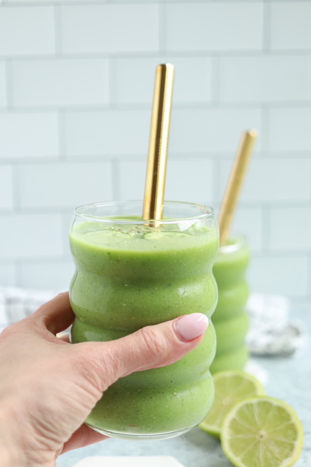 A hand holding a wavy glass of pineapple cucumber smoothie with a gold metal straw; another similar smoothie sits in the background, with sliced lime beside them on a light surface and white tiled wall.