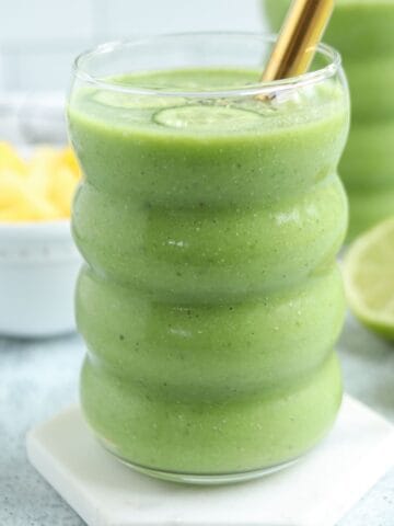 A glass of thick green smoothie with a gold straw, placed on a white coaster. Sliced lime and a bowl of pineapple chunks are in the background, creating a fresh vibe reminiscent of pesto chicken parmesan’s vibrant flavors. The smoothie is in a wavy glass.