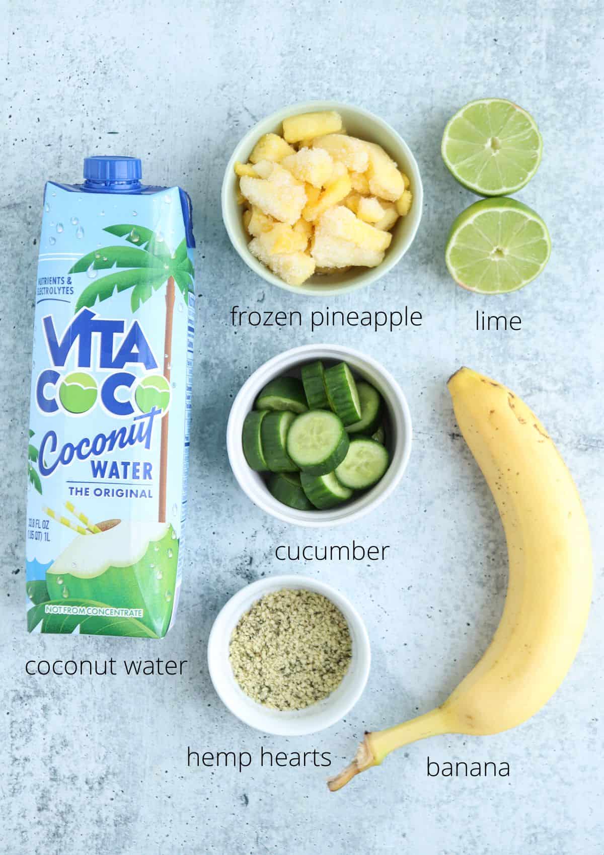 Ingredients for a pineapple cucumber smoothie are arranged on a light surface: a carton of coconut water, frozen pineapple, two lime halves, sliced cucumber, a banana, and hemp hearts, each labeled.