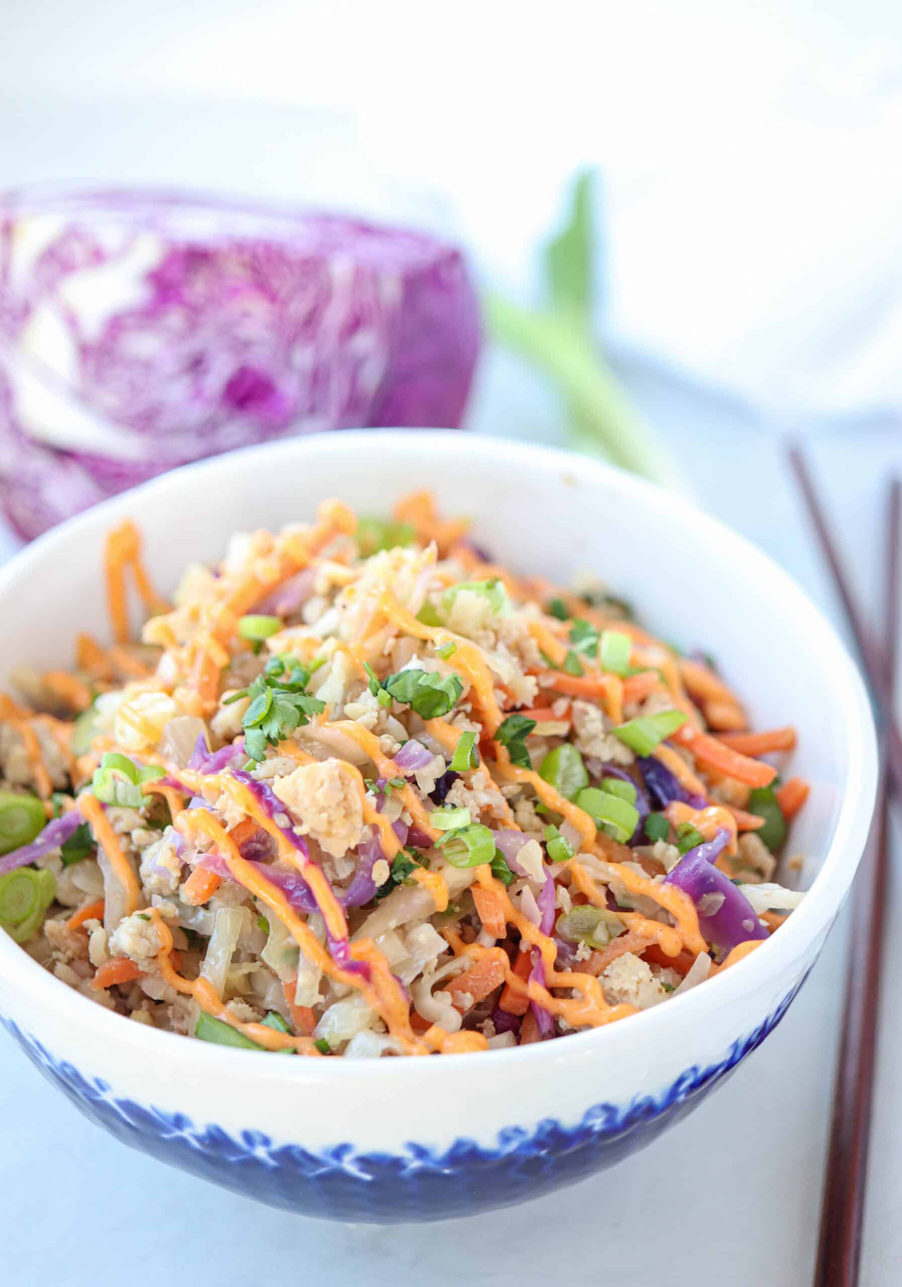 A bowl of colorful stir-fry with shredded carrots, purple cabbage, green onions, and crumbled tofu or chicken, garnished with sesame seeds, sits next to a pair of chopsticks and a piece of purple cabbage in the background.