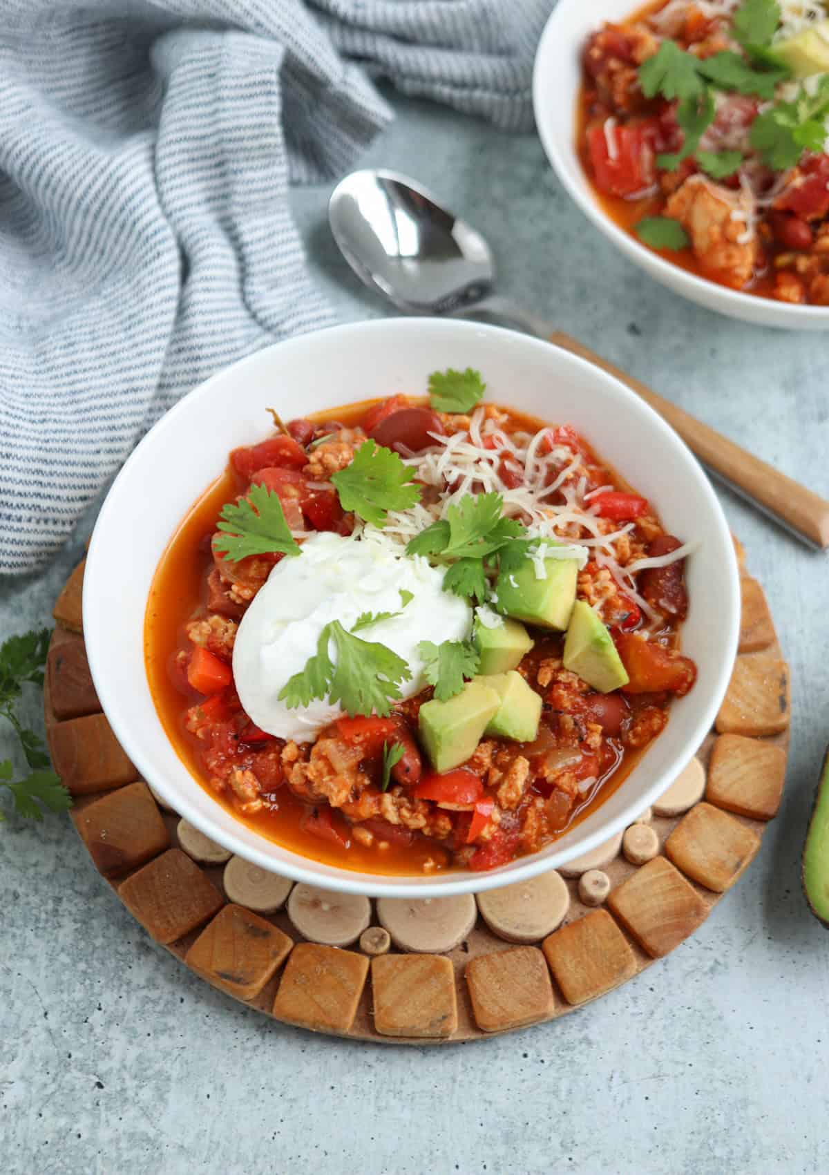 A bowl of chili topped with sour cream, shredded cheese, avocado cubes, and cilantro sits on a wooden trivet, with a spoon and another bowl of chili in the background.