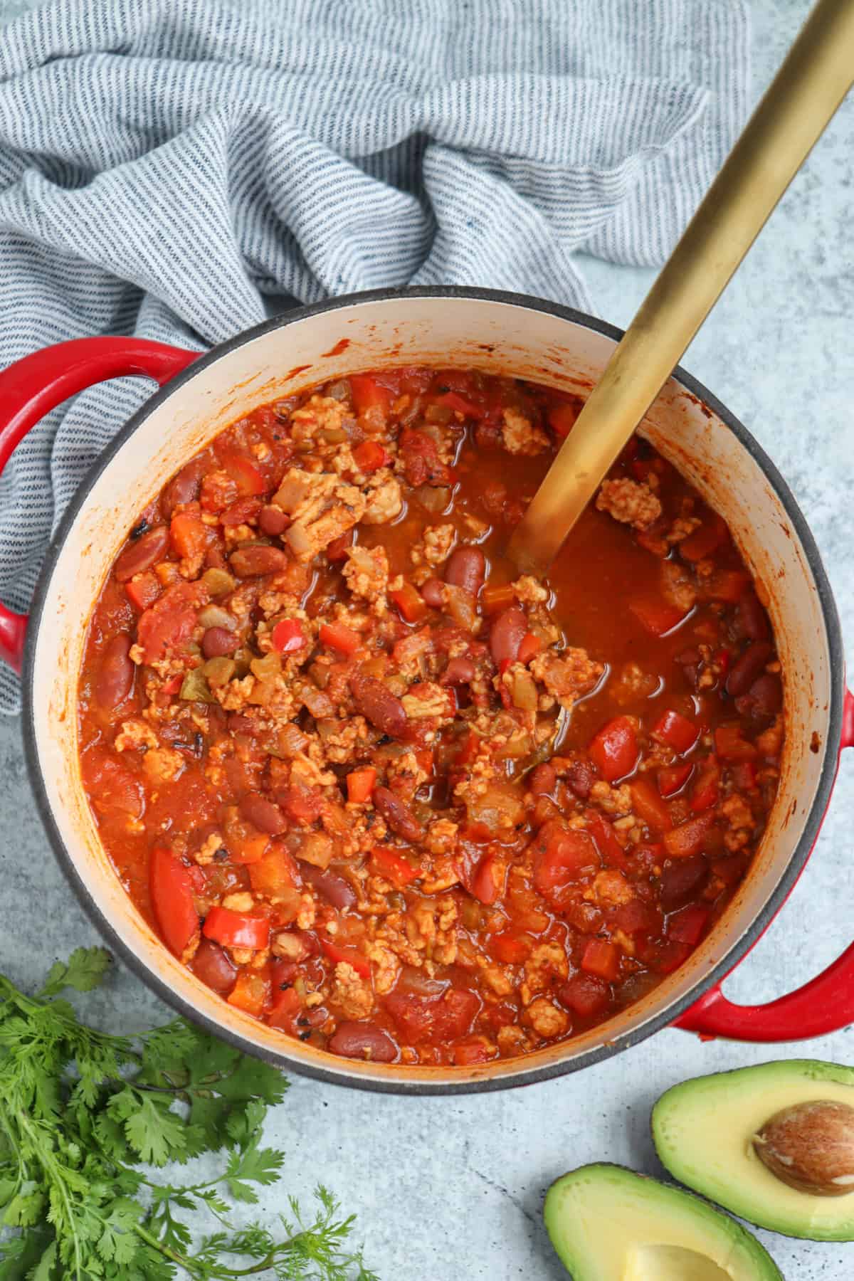 A pot of chili with ground meat, beans, and tomatoes sits on a gray surface, with a gold ladle inside. A striped towel, fresh cilantro, and sliced avocado are nearby—perfect alongside pesto chicken parmesan.
