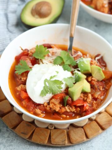 A white bowl of ground chicken chili topped with sour cream, shredded cheese, cilantro, and diced avocado sits on a wooden trivet, with a spoon in the bowl and a half avocado in the background.