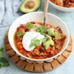 A bowl of chili topped with sour cream, diced avocado, shredded cheese, and cilantro sits on a wooden trivet. A spoon rests in the bowl, with half an avocado in the background—perfect for serving alongside pesto chicken parmesan.