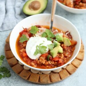 A bowl of chili topped with sour cream, diced avocado, shredded cheese, and cilantro sits on a wooden trivet. A spoon rests in the bowl, with half an avocado in the background—perfect for serving alongside pesto chicken parmesan.