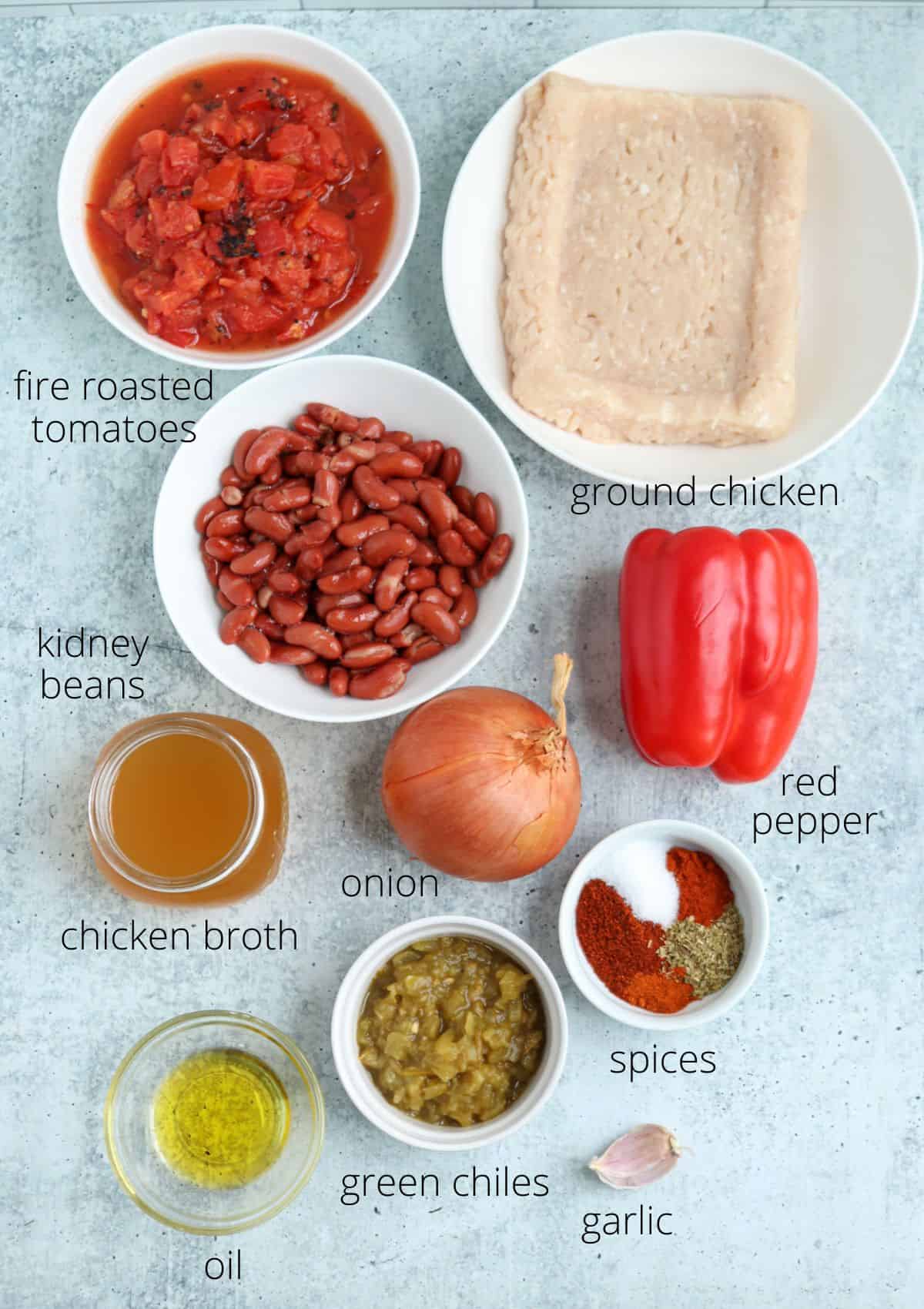 A top-down view of labeled chili ingredients—fire-roasted tomatoes, ground chicken, kidney beans, red pepper, chicken broth, onion, oil, green chiles, spices, and garlic—creates a colorful spread reminiscent of pesto chicken parmesan flavors.