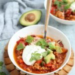 A bowl of ground chicken chili topped with sour cream, shredded cheese, avocado chunks, and cilantro, with a spoon inside. An avocado half and a second bowl of chili are in the background—perfect for pairing with pesto chicken parmesan.