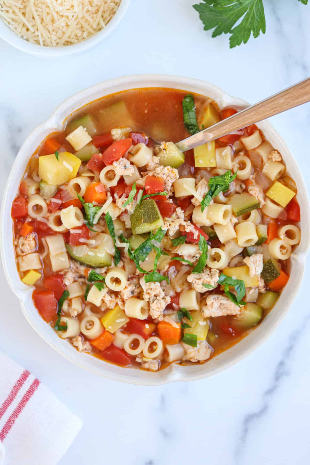 A bowl of vegetable and pasta soup with diced vegetables, pasta, ground meat, and herbs, served with a spoon. A bowl of grated cheese and a parsley sprig are nearby on a white marble surface.