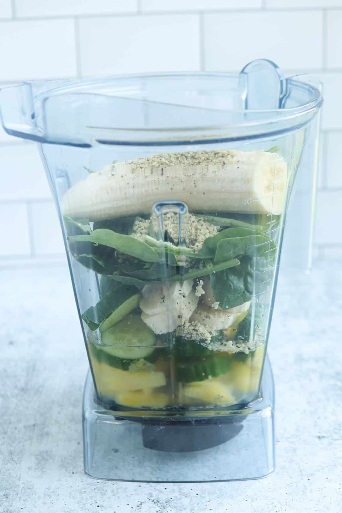 A blender jar filled with a whole banana, leafy greens, pineapple, cucumber slices, seeds, and liquid ingredients for a refreshing pineapple cucumber smoothie sits on a light-colored countertop with a white tiled background.