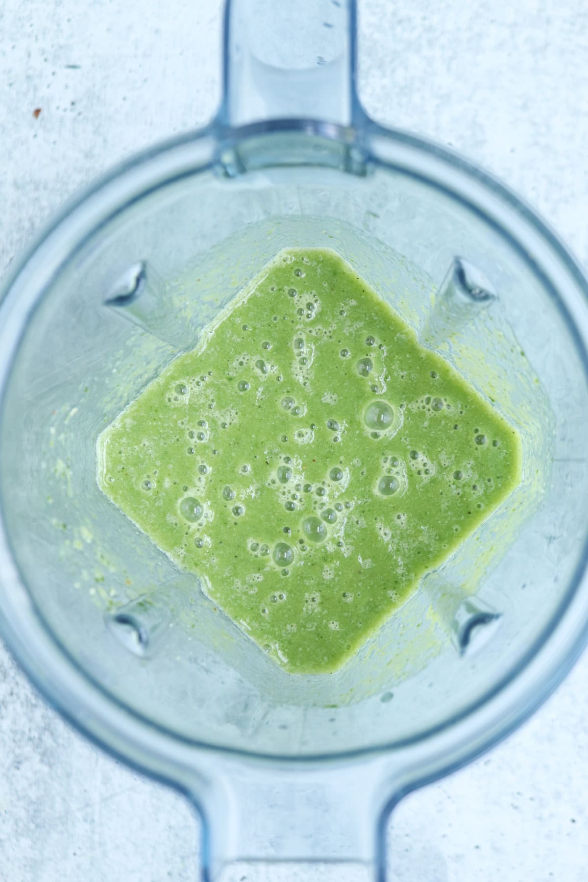 Overhead view of a blender containing a frothy, light green pineapple cucumber smoothie with visible bubbles on its surface. The blender rests on a light, textured background.