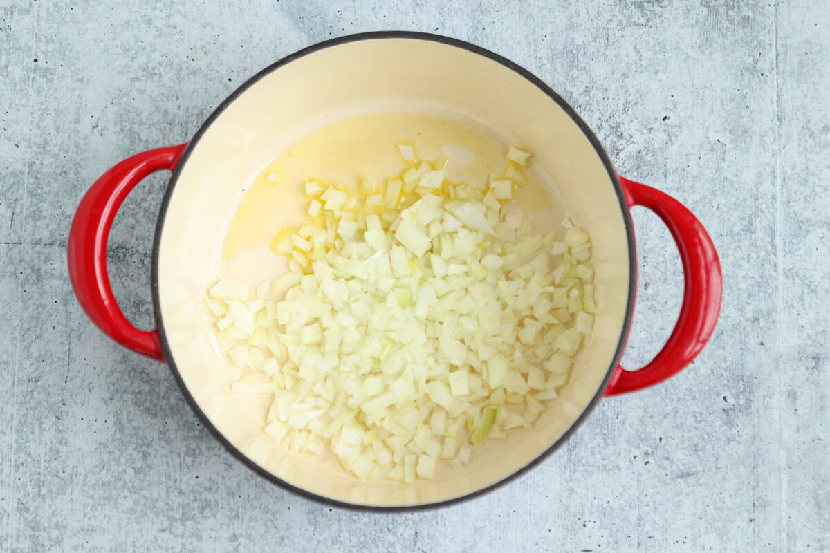 A red-handled pot with chopped onions sautéing in oil, perfect for starting a delicious pesto chicken parmesan, sits on a light gray countertop.