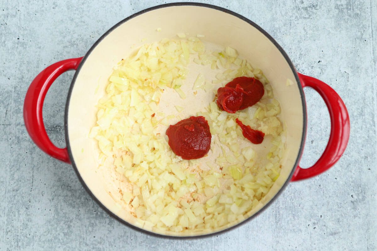 Chopped onions are sautéing in a large cream-colored pot with red handles. Two spoonfuls of tomato paste, perfect for starting a pesto chicken parmesan, rest on the onions, ready to be mixed in. The pot sits on a gray countertop.