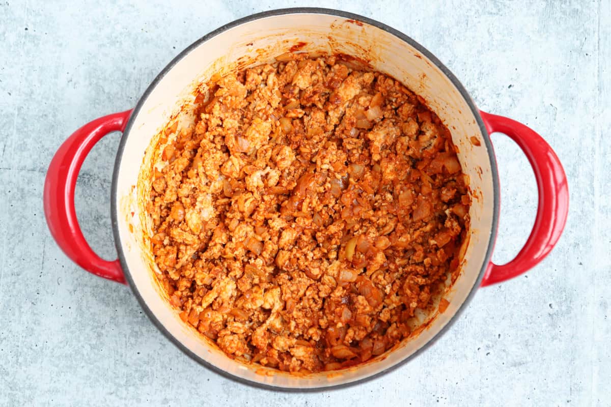 A large red-handled pot filled with cooked ground meat, onions, and tomato sauce—perfect for pairing with pesto chicken parmesan—rests on a light gray countertop.