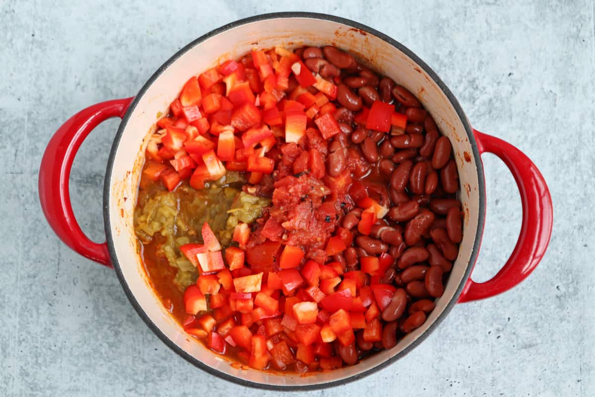 A red-handled pot filled with kidney beans, chopped red bell peppers, diced tomatoes, and green chilies sits on a light gray surface. The uncooked ingredients are arranged in sections, ready for cooking—unlike classic pesto chicken parmesan.