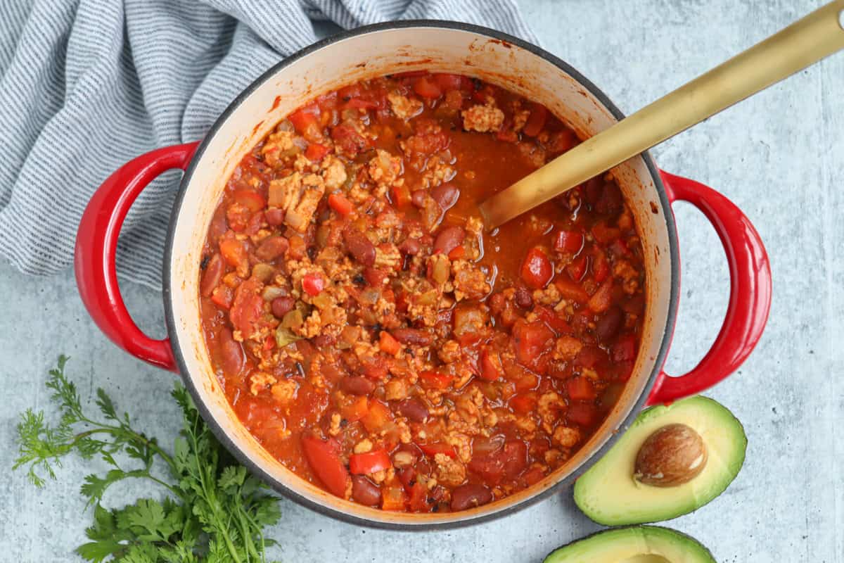A pot of chili with ground meat, beans, and red bell peppers sits on a gray surface. A gold ladle rests inside, and fresh cilantro and a halved avocado are nearby—perfectly cozy alongside pesto chicken parmesan. A striped towel is draped beside the pot.