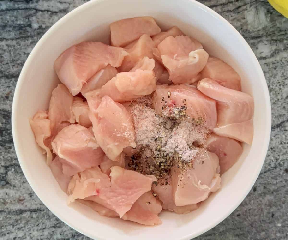 A white bowl filled with raw, cubed chicken pieces sprinkled with salt and black pepper, perfect for preparing pesto chicken parmesan, sits on a gray granite countertop.