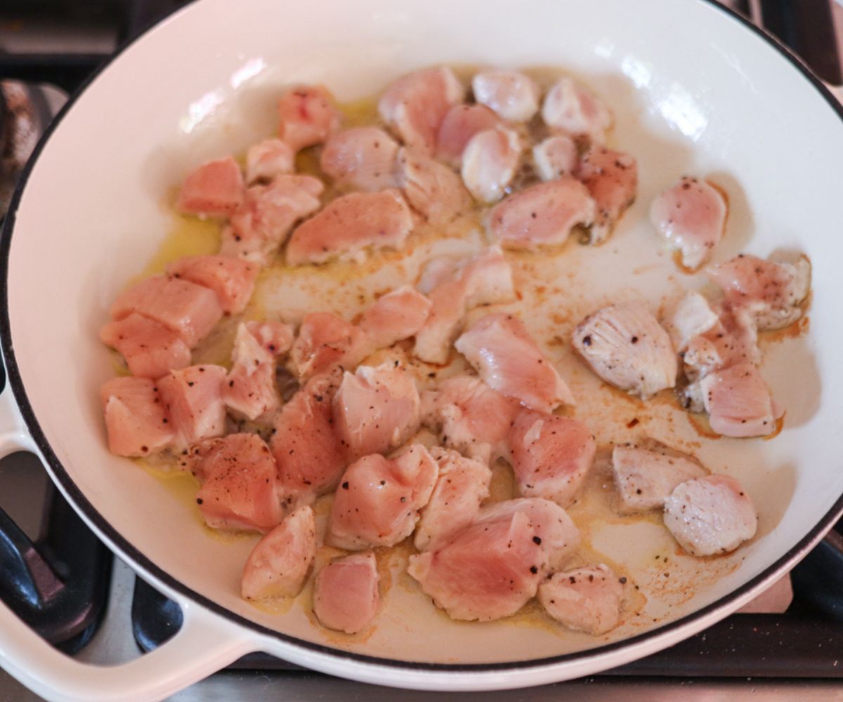 Raw, diced chicken pieces are cooking in a white skillet on a stovetop, with some seasoning and oil visible—perfect for starting a flavorful pesto chicken parmesan.