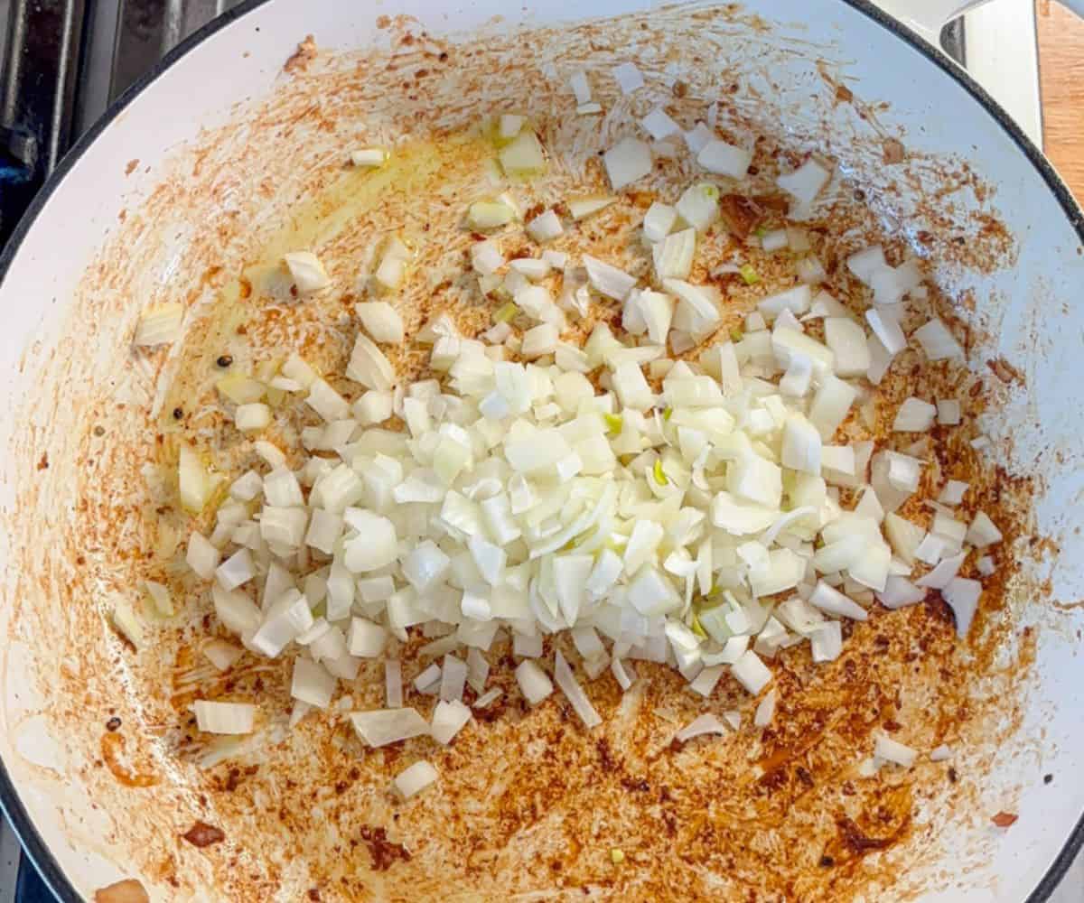 Chopped onions piled in the center of a large white pot with browned seasoning and residue on the sides, hinting at the delicious beginnings of a pesto chicken parmesan cooking process.