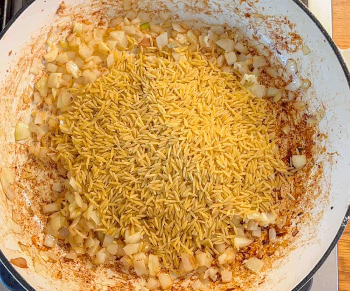 A pot with diced onions being sautéed, mixed with uncooked rice and browned bits stuck to the sides, sets the foundation for a delicious pesto chicken parmesan recipe.