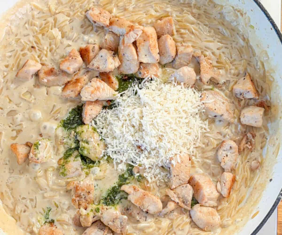 A skillet filled with creamy orzo pasta, pesto chicken parmesan, and a drizzle of green pesto sauce, all partially mixed together and topped with grated cheese.
