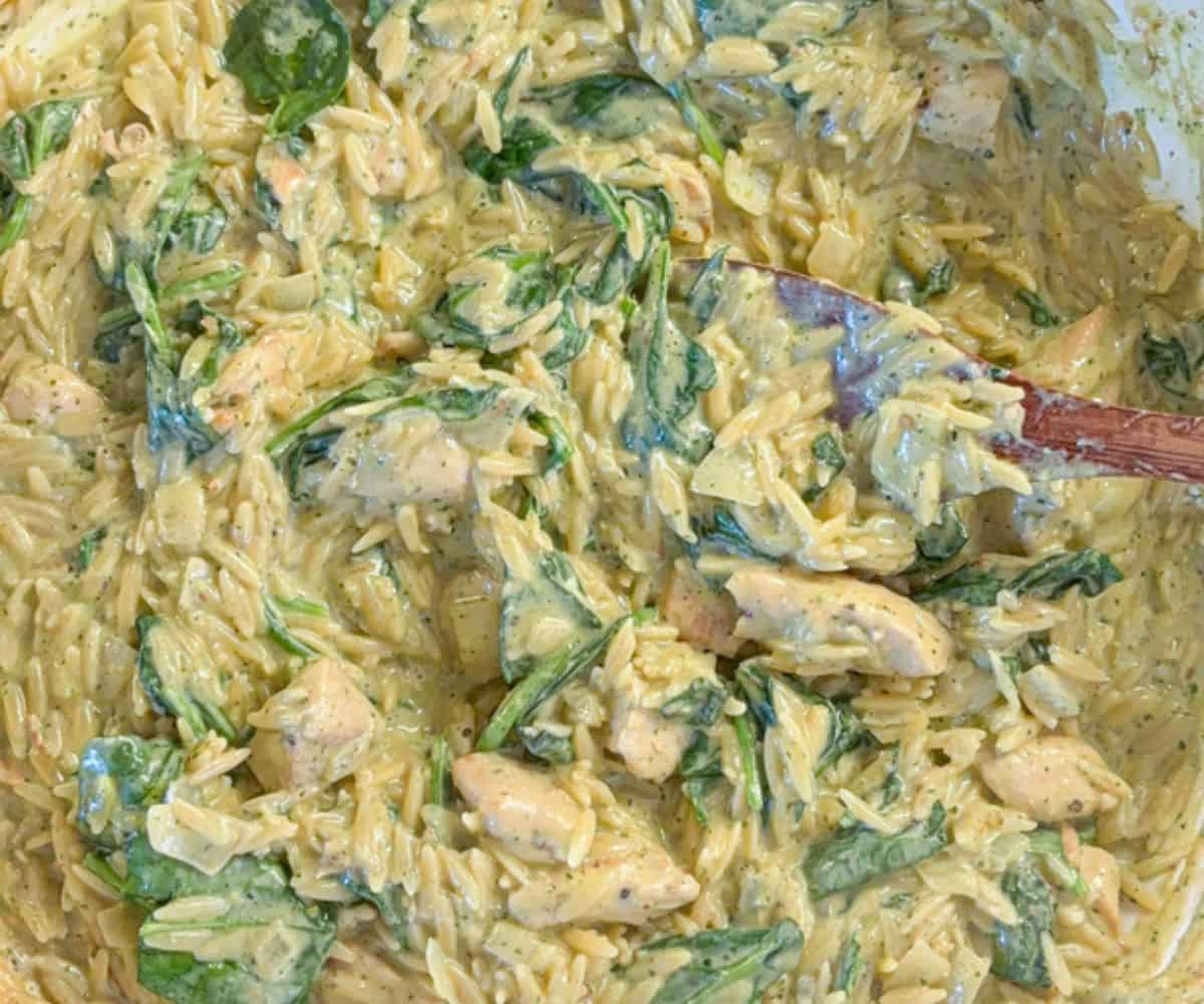 Creamy orzo pasta mixed with pesto chicken parmesan and fresh spinach, all coated in a thick, seasoned sauce. A wooden spoon is partially visible, stirring the ingredients together.