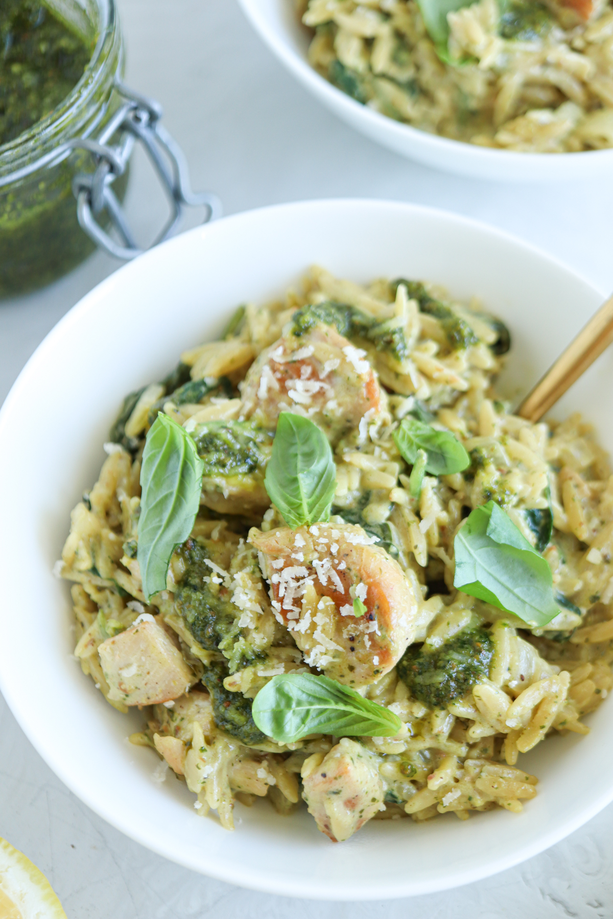 A white bowl filled with creamy orzo pasta, pesto chicken parmesan, shrimp, spinach, and garnished with fresh basil leaves and grated cheese. A fork rests inside the bowl.