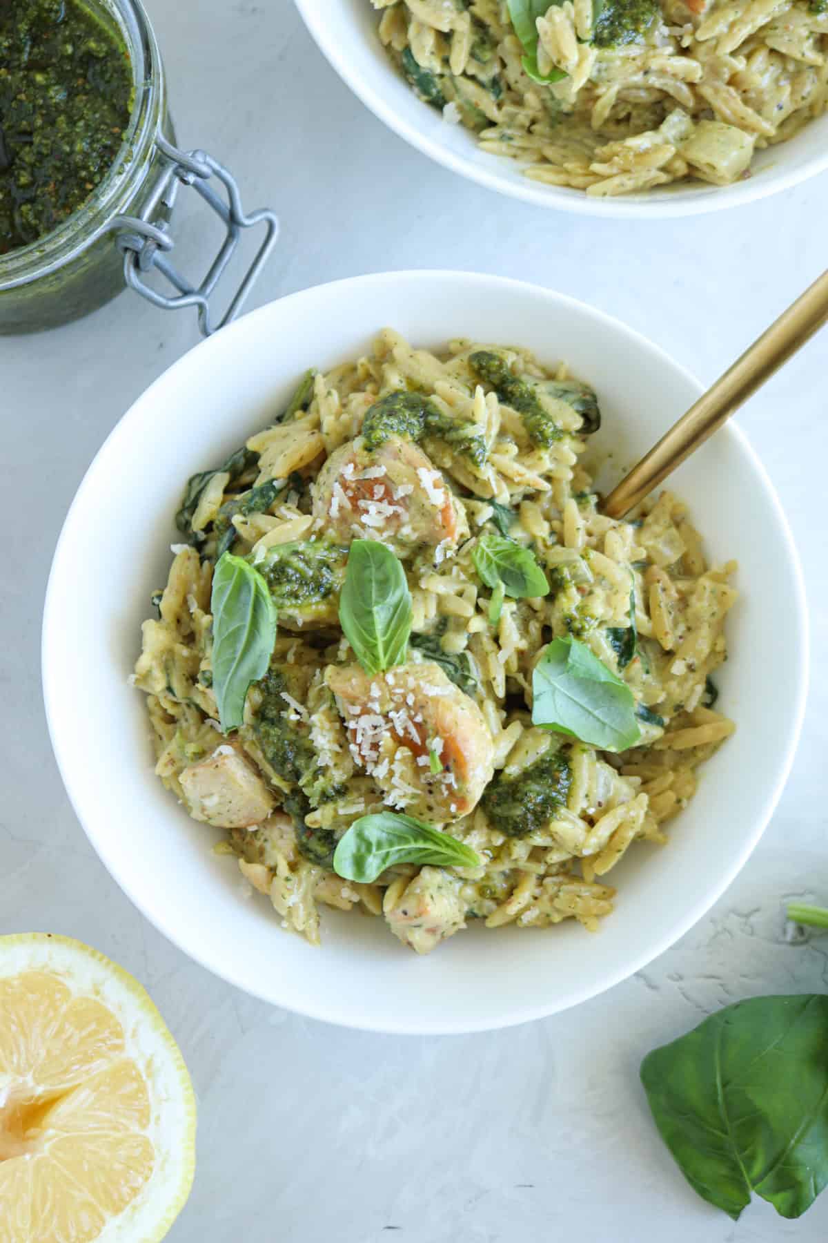 A white bowl filled with pesto chicken orzo, fresh basil leaves, and grated cheese. A gold fork rests in the bowl. A lemon half and a jar of pesto are nearby on a light surface.