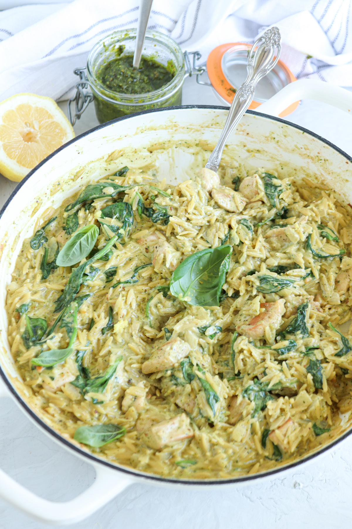 A creamy pesto chicken orzo and spinach dish in a white pot, garnished with fresh basil leaves, with a jar of green pesto, a spoon, and a halved lemon beside it on a light-colored surface.