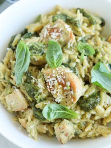 A bowl of orzo pasta with tender pesto chicken pieces, topped with fresh basil leaves and a sprinkle of grated Parmesan cheese.